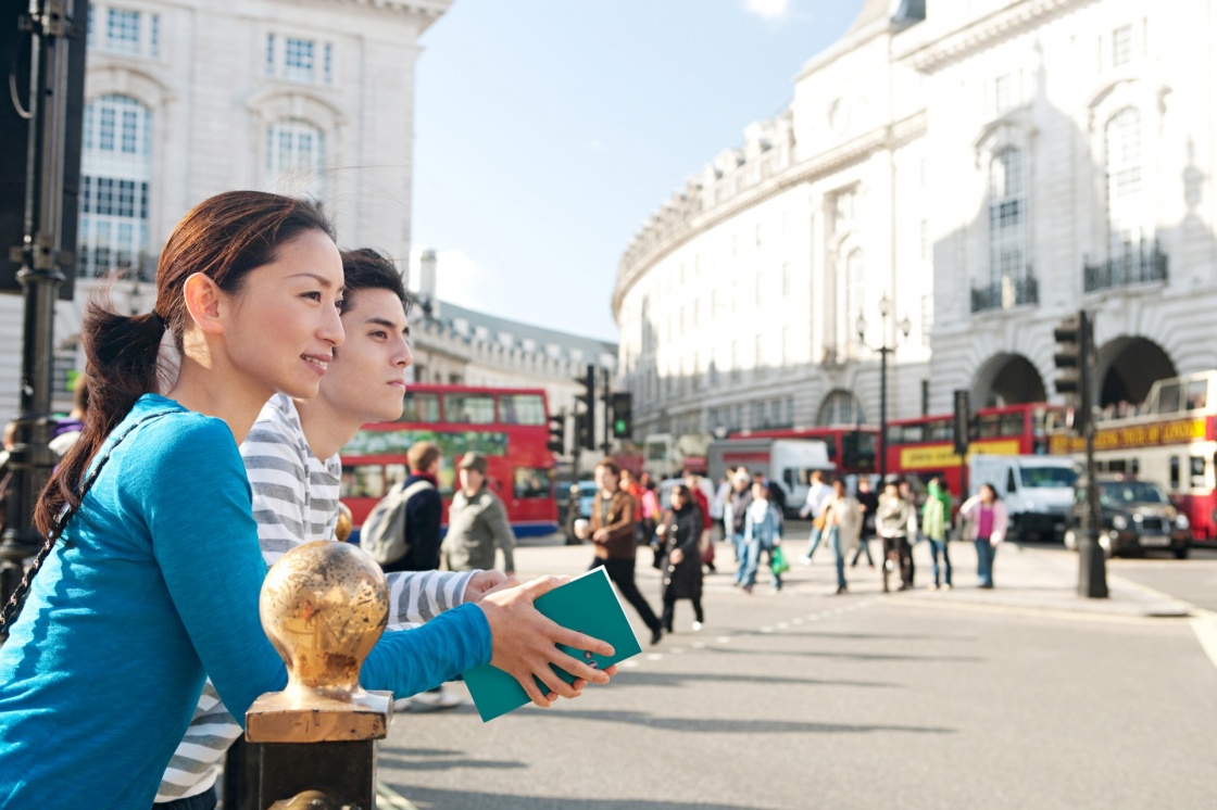 'Side portrait view of a young Japanese tourist couple in Piccadilly Circus landmark street with red buses, while visiting the city of London on holiday, smiling during a sunny day outdoors.' - Λονδίνο